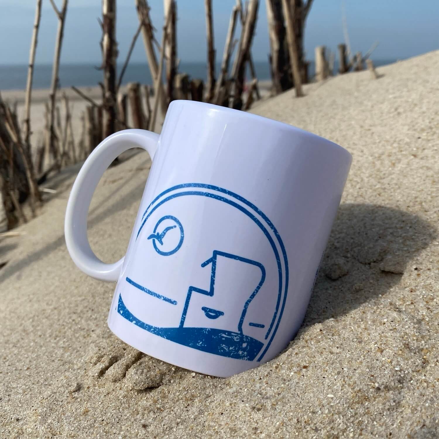 Weißer Syltbecher liegt am Strand auf Sylt. Im Hintergrund ist der Strand zu sehen. Der Becher ist in blau bedruckt mit dem Begriff Sylt, einem Sylter Nordsee Strandkorb sowie einer Möwe die durch die Sonne fliegt.