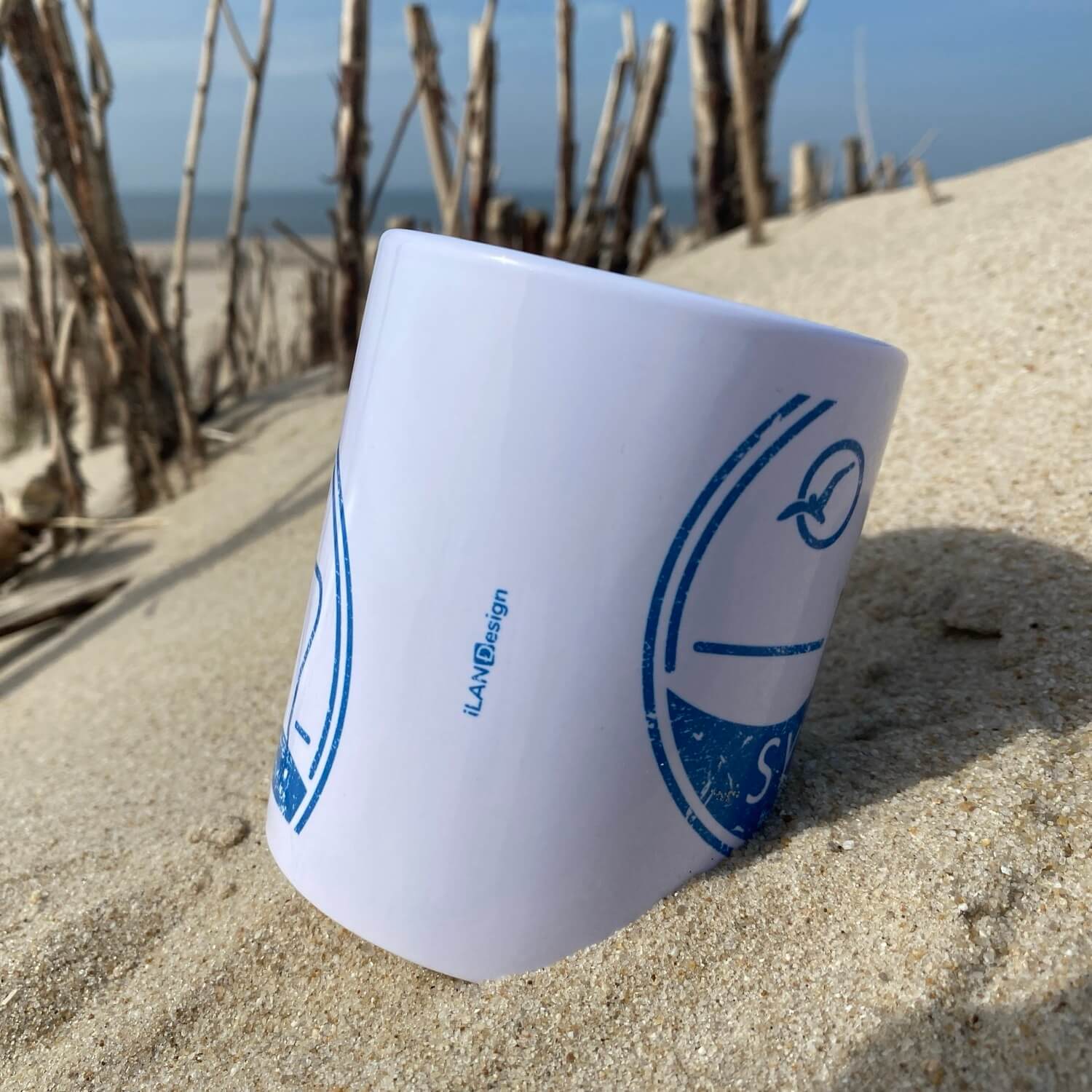 Weißer Becher liegt am Strand auf Sylt. Im Hintergrund ist das blaue Meer zu sehen. Der Becher ist in blau bedruckt mit dem Begriff Sylt, einem  Strandkorb sowie einer Möwe die durch die Sonne fliegt.
