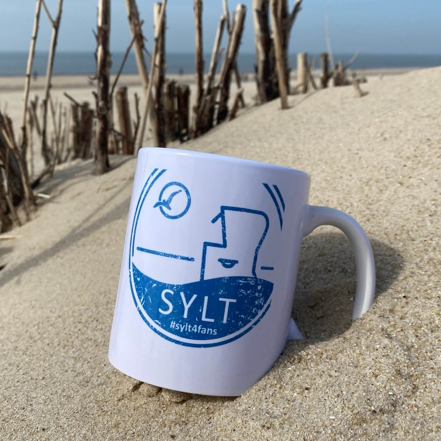Weißer Keramikbecher liegt am Strand auf Sylt. Im Hintergrund ist das Meer zu sehen. Der Becher ist in blau bedruckt mit dem Begriff Sylt, einem Sylter Nordsee Strandkorb sowie einer Möwe die durch die Sonne fliegt.