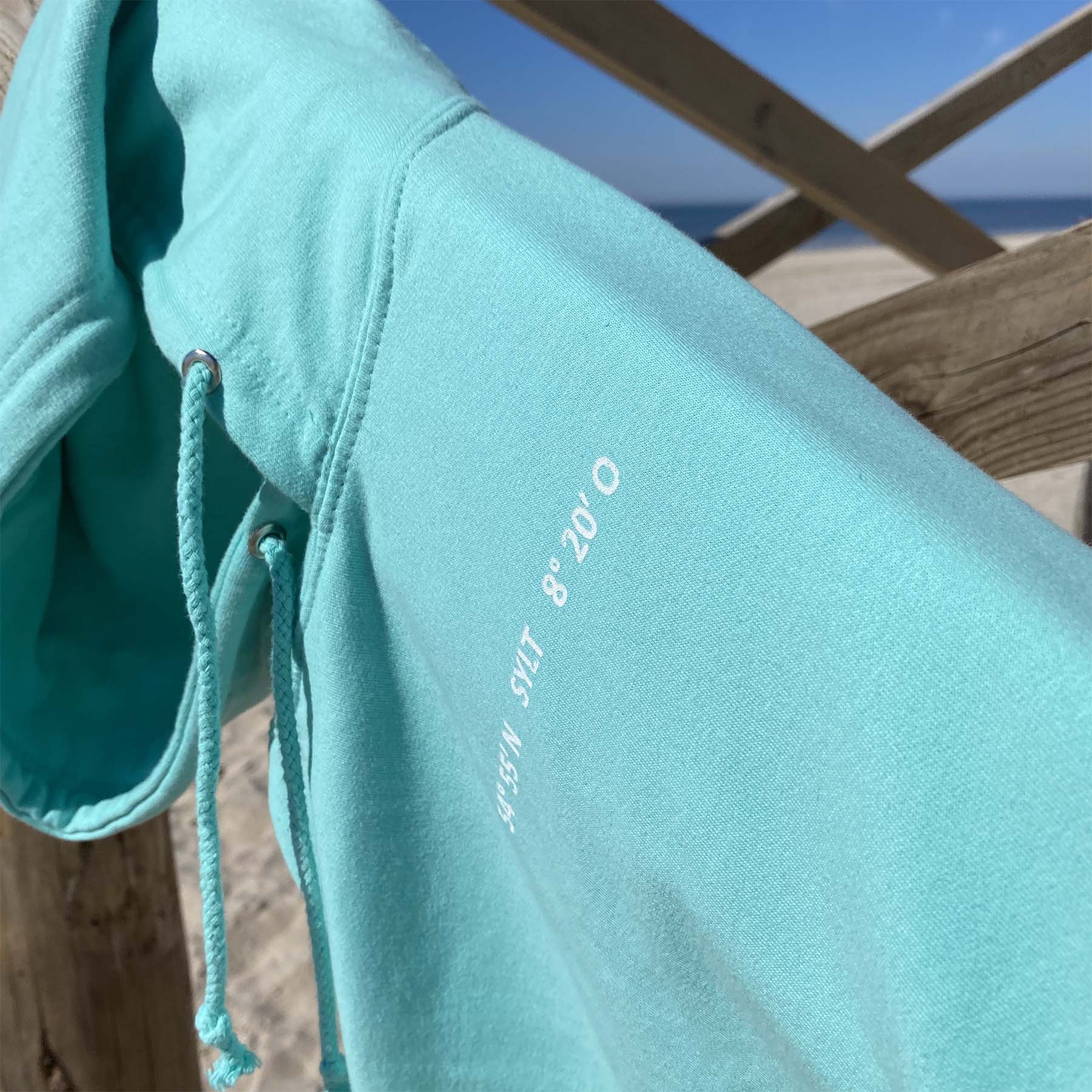 Sylt Hoodie im Aqua-Ton mit Fokus auf den weißen Schriftzug Sylt und Koordinaten