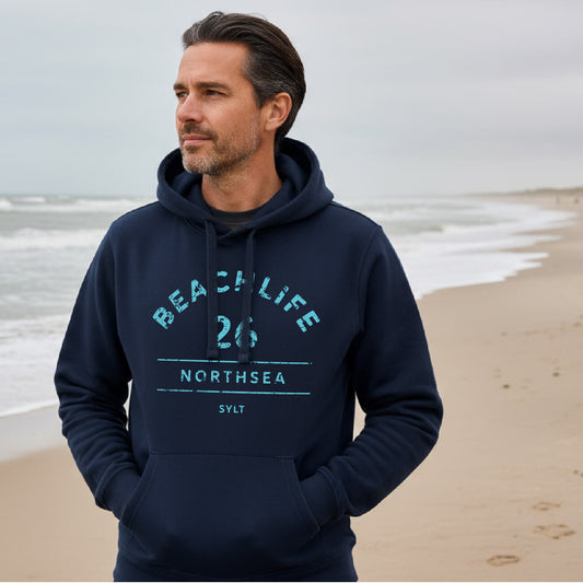 Mann steht mit Sylt Hoodie Beachlife 26 in dunkelblau am Nordseestrand. Der Aufdruck ist in einem frischen türkis.