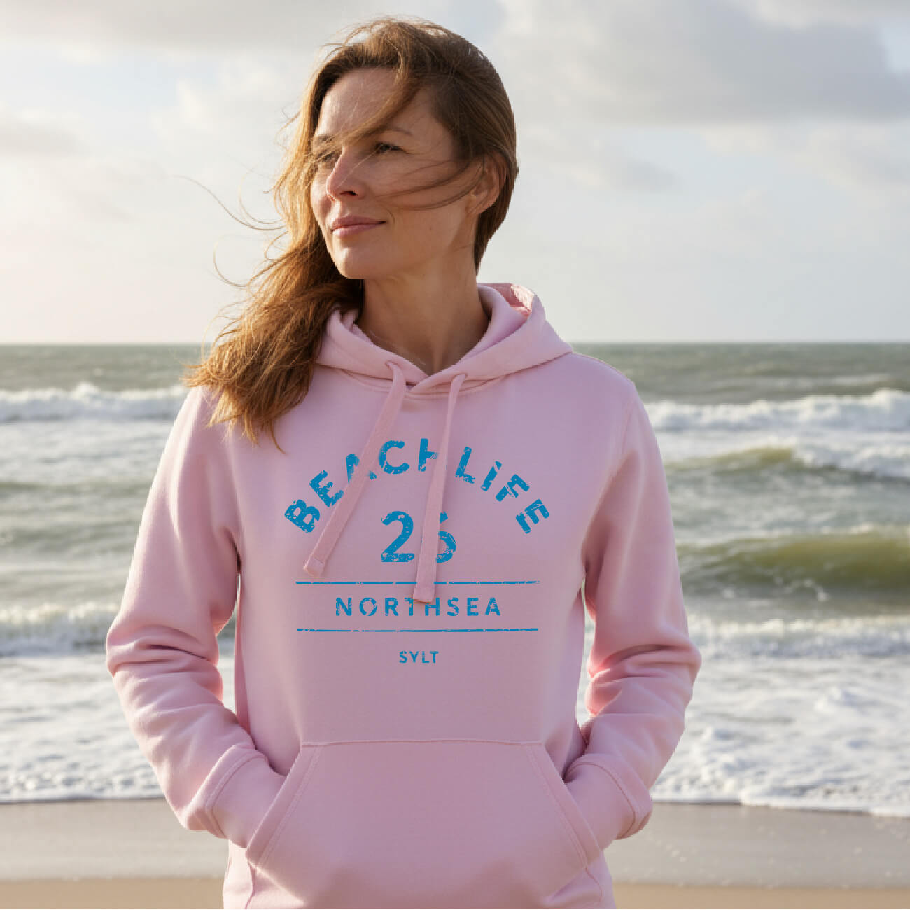 Frau im rosa Beachlife 16 Sylt Hoodie am Strand. Im Hintergrund sieht man die raue Sylter Nordsee.