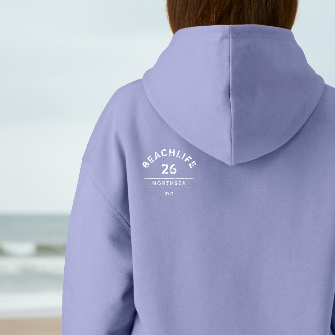 Rückenansicht des violeten BEACHLIFE 26 Hoodie mit dezentralem Print in natürlicher Strandatmosphäre.
