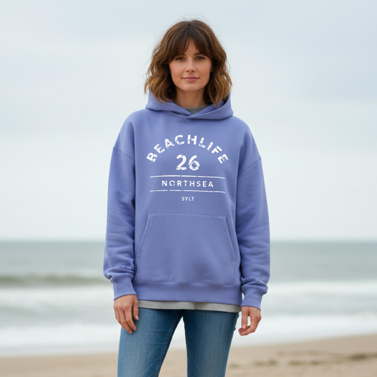 Braunhaarige Frau in violetem Hoodie mit "BEACHLIFE 26 NORTHSEA SYLT" Aufdruck in entspannter Strandszenerie.