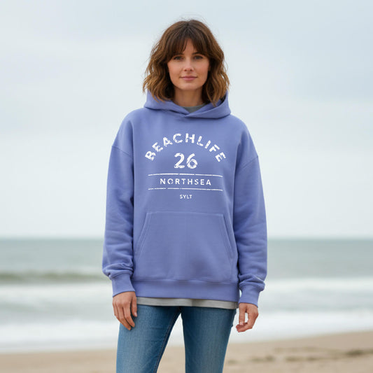 Braunhaarige Frau in violetem Hoodie mit "BEACHLIFE 26 NORTHSEA SYLT" Aufdruck in entspannter Strandszenerie.