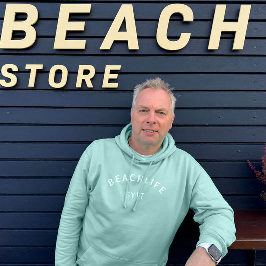 Mann steht mit einem aqua farbigem Sylt Hoodie vor einem schwarzen Strandhaus. Vorne auf dem Hoodie ist der Begriff Beachlife leicht gerundet aufgedruckt. Darunter steht mittig in weiß Sylt.