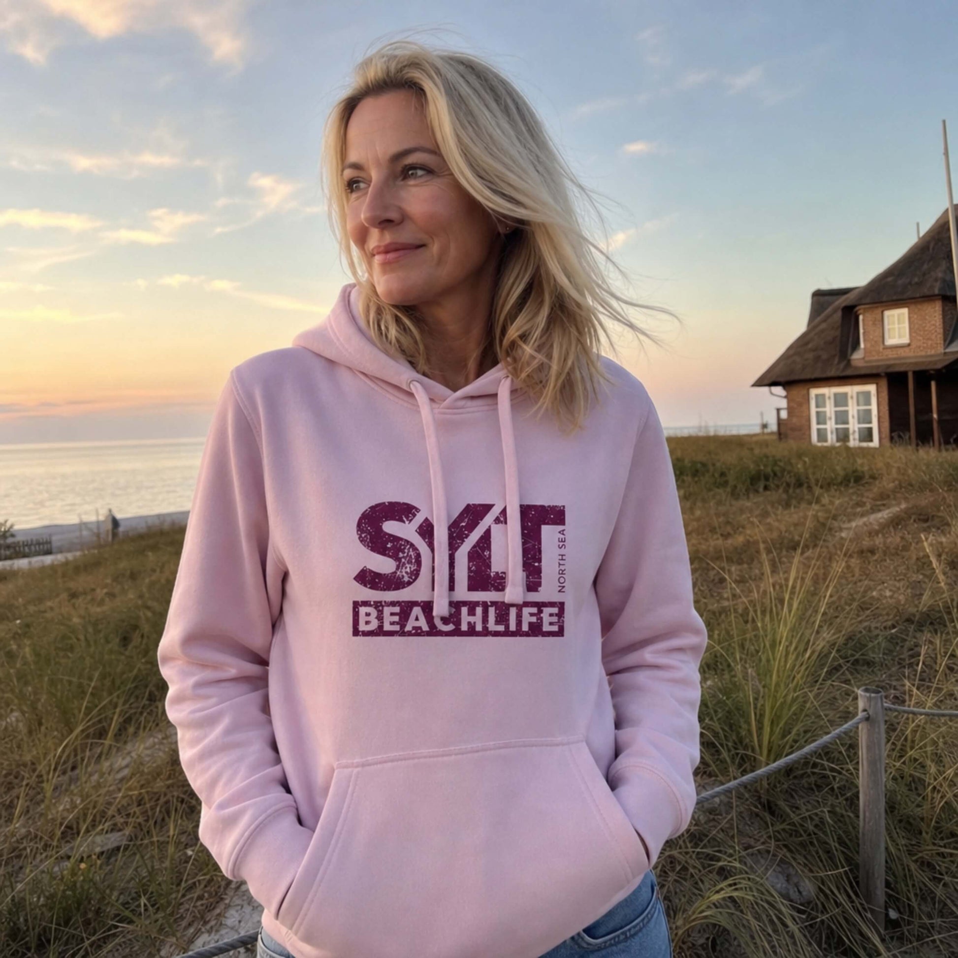 Frau in rosanem Hoodie mit rosanem 'SYLT NORTH SEA BEACHLIFE' Logo, steht auf Sylt.