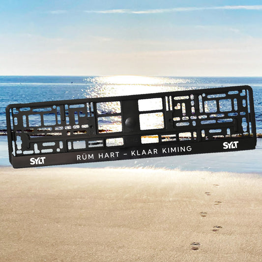 Sylt Kennzeichenhalter mit modernem Sylt-Schriftzug links und rechts. In der Mitte steht der friesische Spruch "Rüm hart – Klaar Kiming". Im Hintergrund des Nummernschildhalters sieht man die Sylter Nordsee mit Spiegelung des Sonnenscheins.