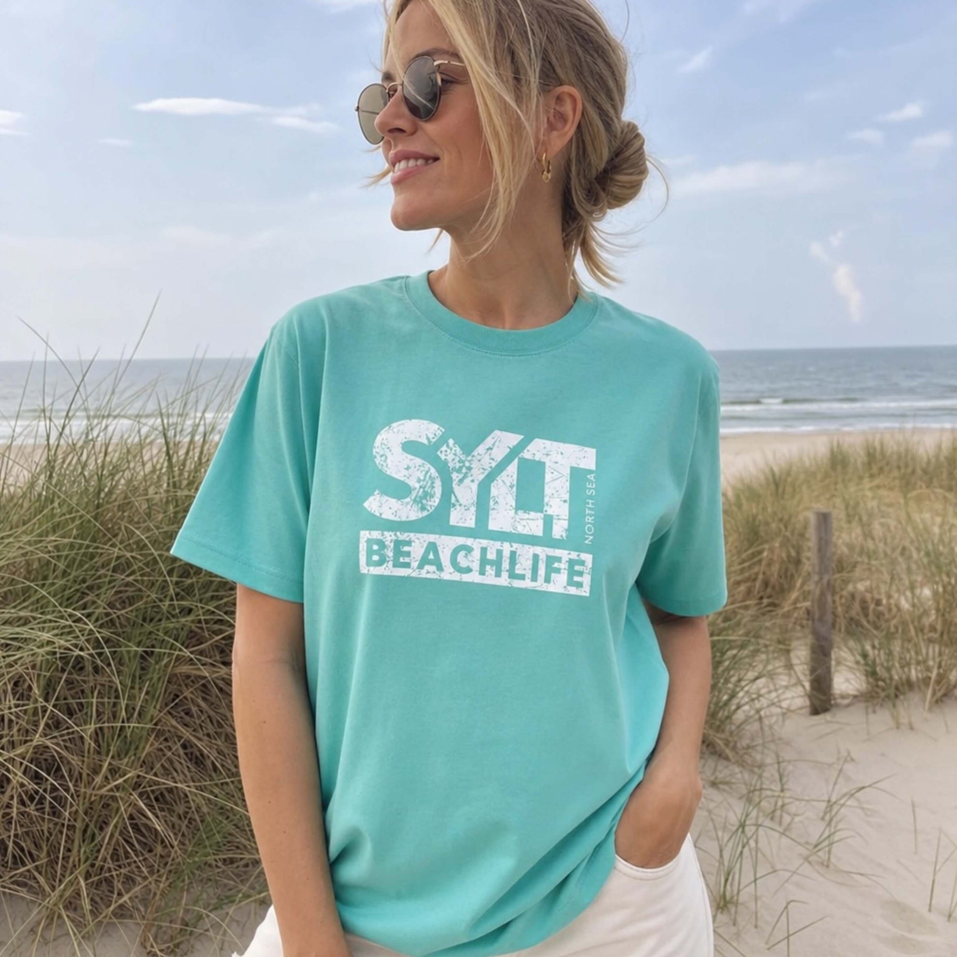 Frau in aqua farbenden Sylt T-Shirt steht auf sylt. Im Hintergrund sieht man Dünen, Strandkörbe am Strand und die Nordsee.