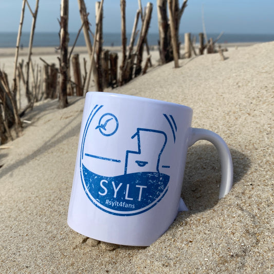 Weißer Keramikbecher liegt am Strand auf Sylt. Im Hintergrund ist das Meer zu sehen. Der Becher ist in blau bedruckt mit dem Begriff Sylt, einem Sylter Nordsee Strandkorb sowie einer Möwe die durch die Sonne fliegt.