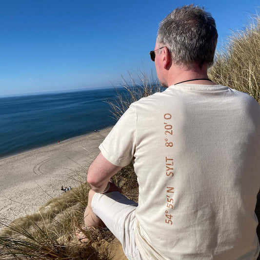 Mann sitzt am Roten Kliff auf Sylt und schaut hinunter auf die Nordsee und den Strand. Der Mann ist von hinten zu sehen. Er trägt ein beiges T-Shirt aus Biobaumwolle.  Von oben nach unten sind auf der linken Seite des Shirts die Koordinaten von Sylt sowie der Begriff Sylt in einem Kupferton aufgedruckt.