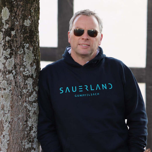 Mann mit dunkelblauem Hoodie steht an einen Baum angelehnt vor einem Fachwerkhaus. Der dunkelblaue Hoodie ist mit dem new Sauerland Logo bedruckt sowie mit dem Dorfnamen Gumpfelbach.