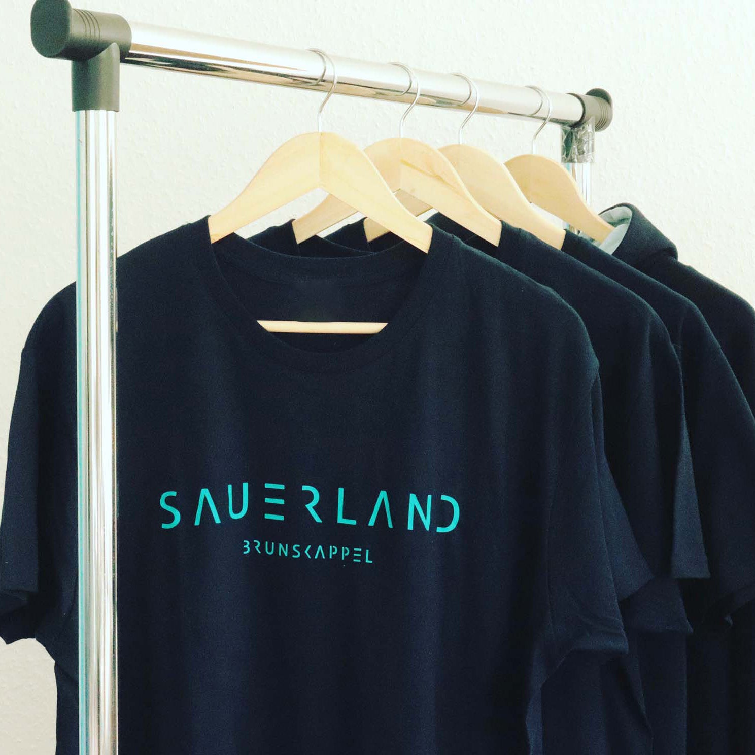 Kleiderstange mit dunkelblauen T-Shirts. In türkis ist der new Sauerland-Schriftzug aufgedruckt. Darunter, ebenfalls in türkis, ist der Dorfname Brunskappel aufgedruckt. Jeder Sauerländer Dorfname kann auf Wunsch aufgedruckt werden.