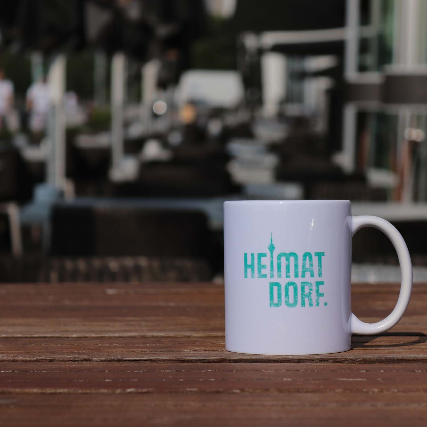 Weißer Kaffebecher mit Düsseldorf HeimatDorf Logo in türkis aufgedruckt, steht auf einem Tisch in der Außengastronomie im Düsseldorfer Medienhafen. Der Aufdruck ist mit einem abgewetzten Grunge-Effekt versehen. Das i von HeimatDorf ist durch den Rheinturm dargestellt.