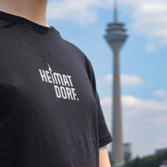 Junger Mann steht in einem schwarzen T-Shirt mit dem HeimatDorf Logo aufgedruckt vor dem Düsseldorfer Rheinturm.