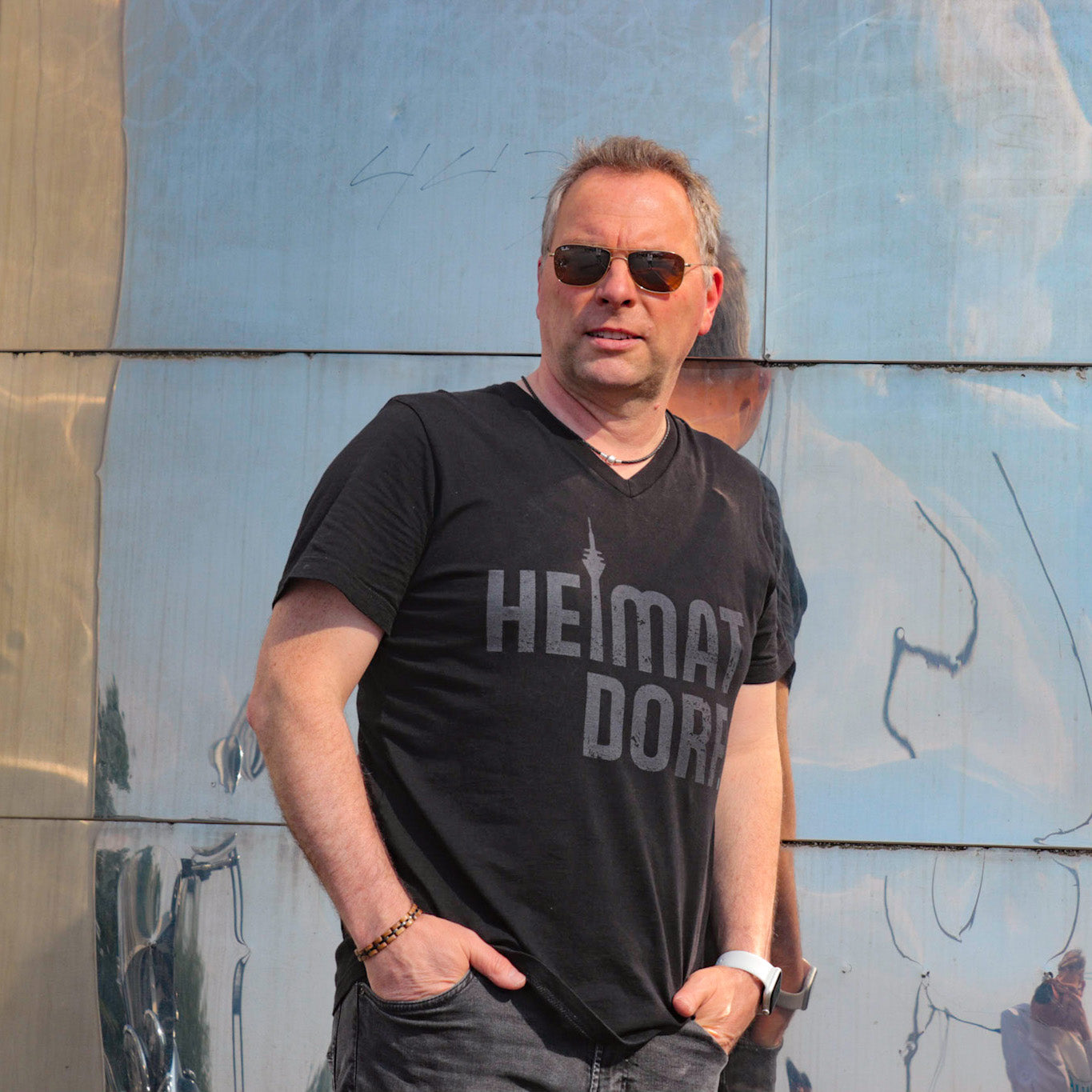 Mann lehnt im Düsseldorfer Medienhafen an einem der Gehry-Häuser. Der Mann trägt ein schwarzes T-Shirt mit V-Ausschnitt. Das Shirt ist in groß mit einem grauen Heimatdorf Schriftzug bedruckt. Der Buchstabe i in Heimatdorf ist durch den stilisierten Rheinturm dargestellt.