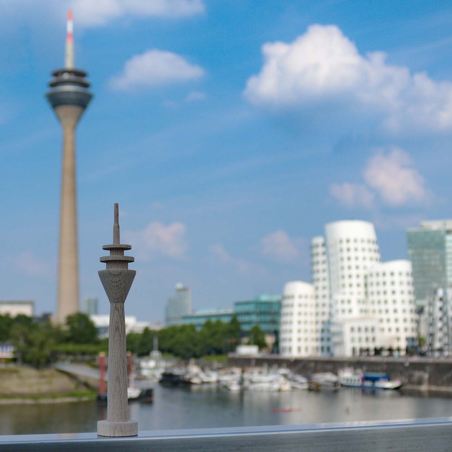 Kleiner Holz-Rheinturm steht im Düsseldorfer Medienhafen auf einem Geländer. Im Hintergrund sieht man den echten Rheinturm, den Hafen sowie die Gehry-Häuser.