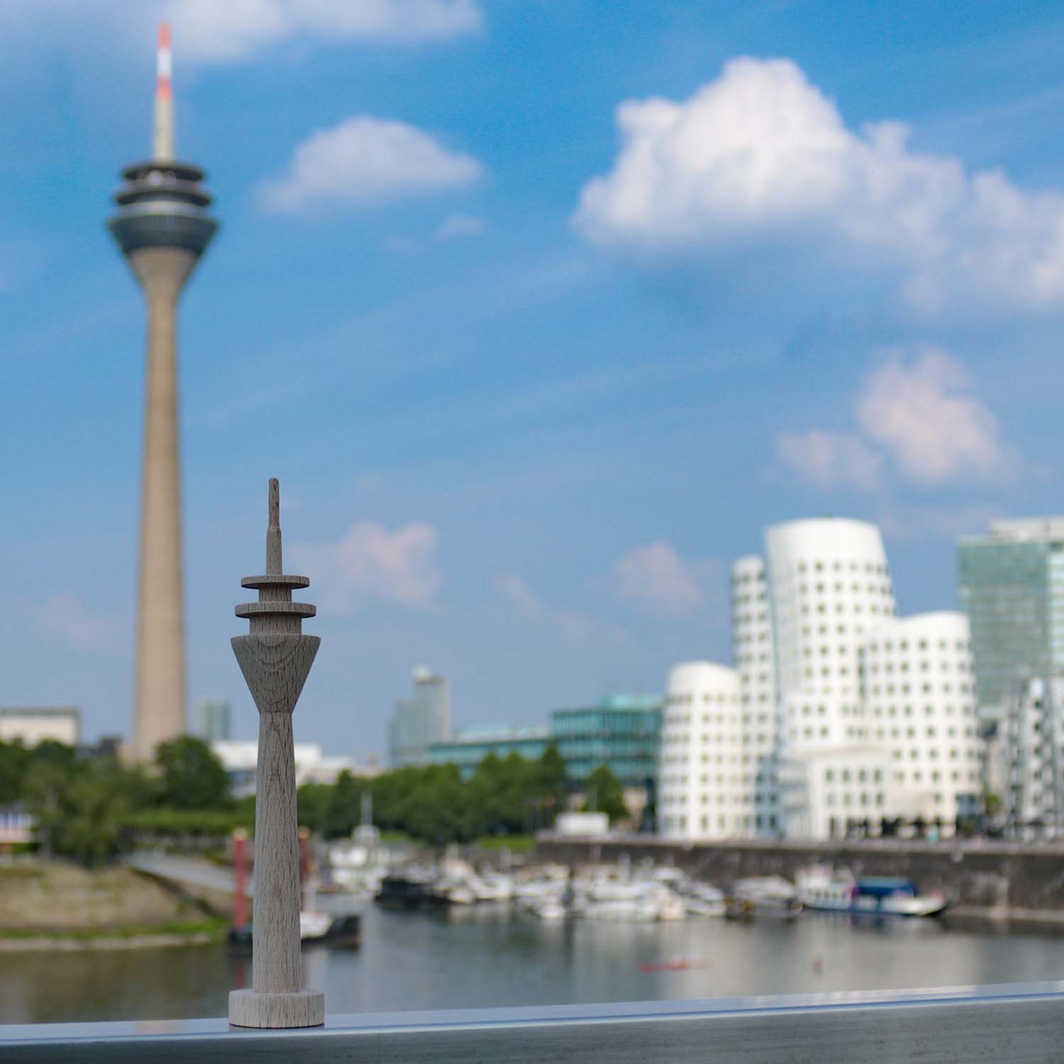 Kleiner Holz-Rheinturm steht im Düsseldorfer Medienhafen auf einem Geländer. Im Hintergrund sieht man den echten Rheinturm, den Hafen sowie die Gehry-Häuser.