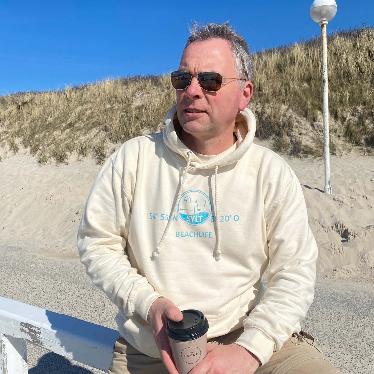 Mann sitzt mit Coffee to go Becher am Sylter Strand und hat einen Hellbeigen Hoodie an. Der Hoodie ist in türkis mit den Koordinaten von Sylt, dem Begriff Sylt sowie einer Grafik von einem Sylter Nordsee-Strandkorb bedruckt. Darunter ist der begriff Beschlief aufgedruckt.