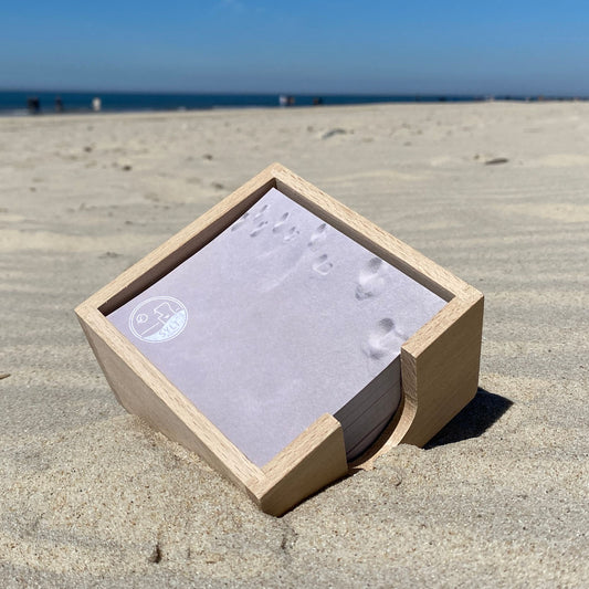 Holz Zettelbox mit quadratischen Notizzetteln liegt auf Sylt im Sand. Im Hintergrund sieht das Meer und blauen Himmel. Die Notizzettel sind mit einem hellen Sandmotiv mit Fußspuren bedruckt sowie einem runden Logo mit Sylt-Schriftzug und Nordessestrandkorb. 