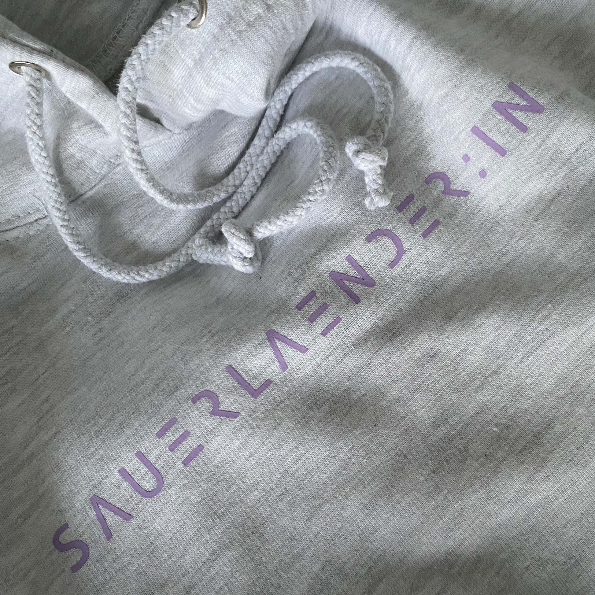 Auf dem Foto sieht man einen Ausschnitt von einem hellgrauen Damen Hoodie mit lavendelfarbiger Beschriftung. Man liest den geänderten Begriff Sauerlaender:in im modernen Sauerland Schriftzug.