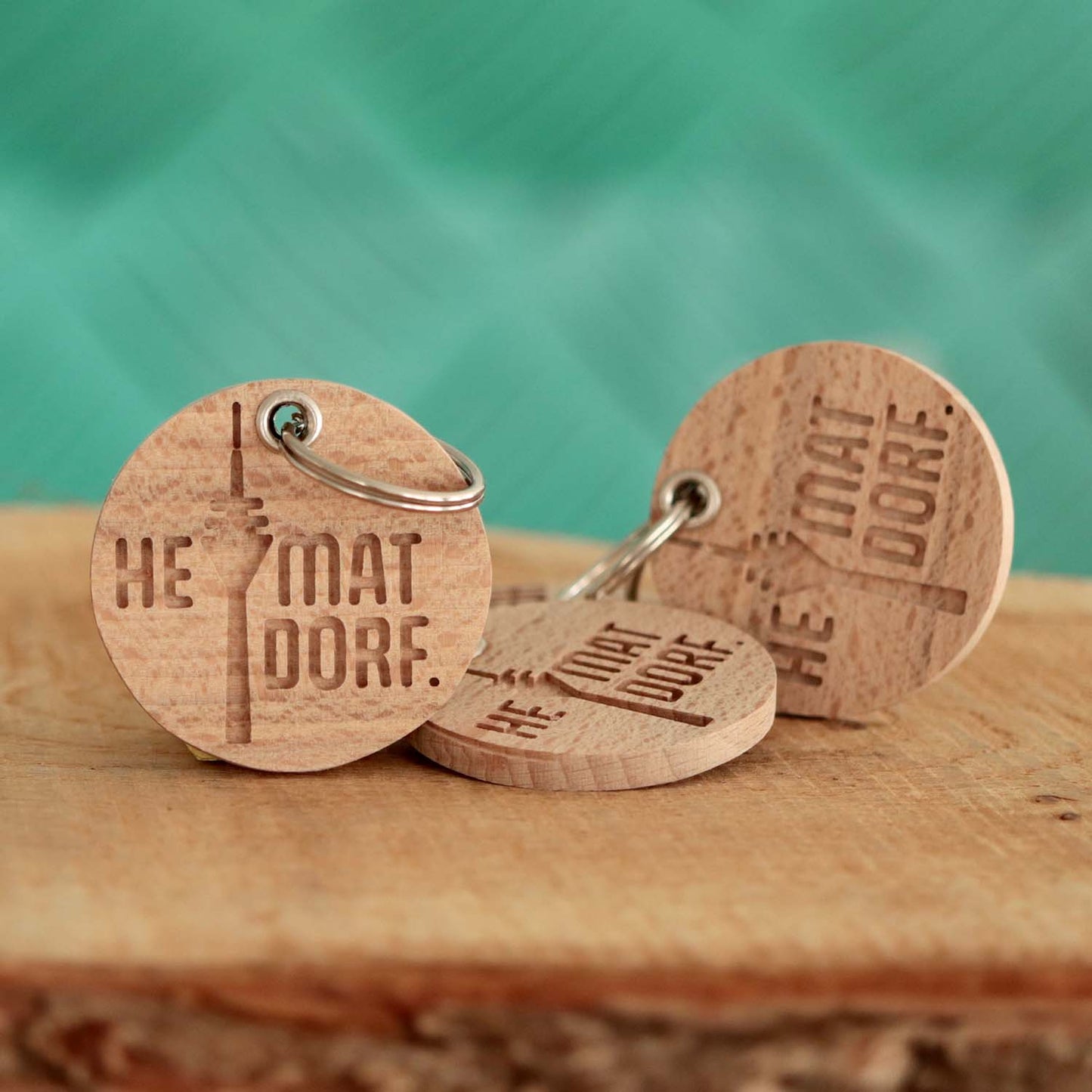 Drei rund Holzschlüsselanhänger mit dem Düsseldorf HeimatDorf Logo liegen auf einem Holzbrett. Die Schlüsselanhänger haben ein verstärktes Loch für einen silbernen Schlüsselring.