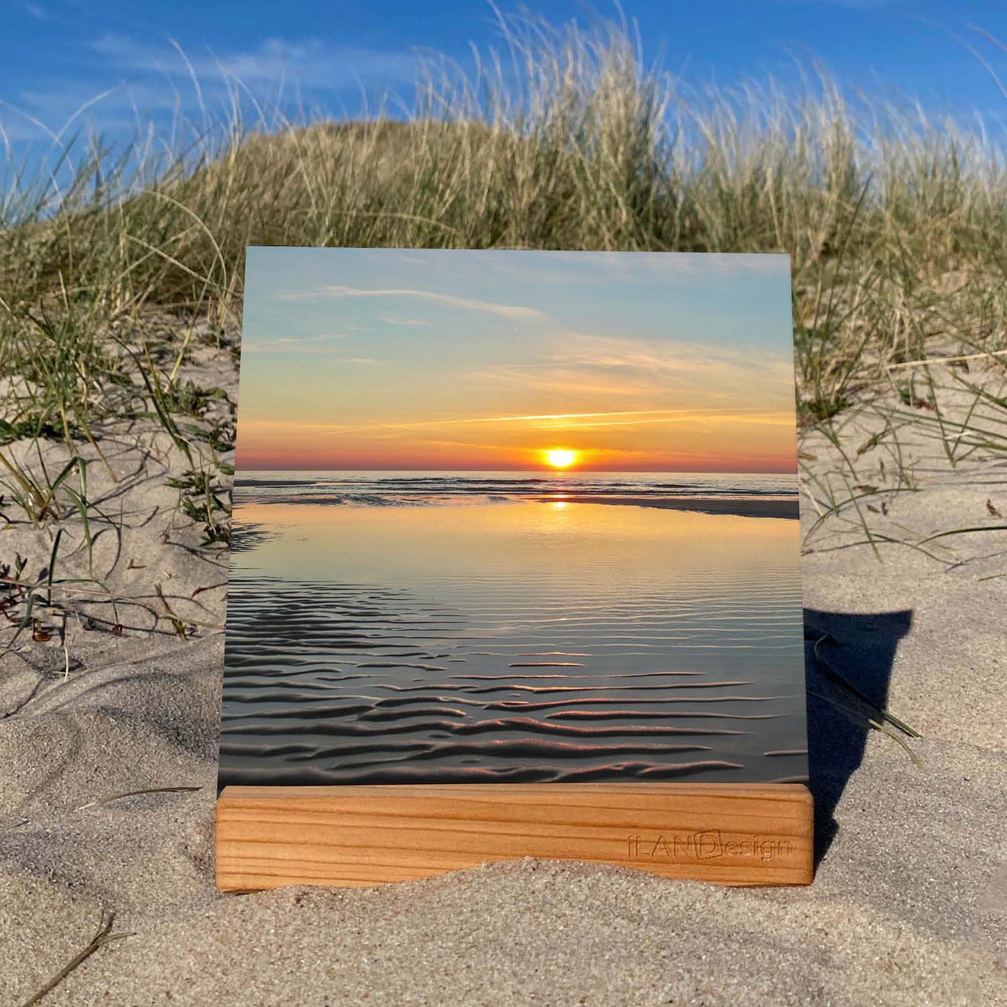 Auf dem Foto ist ein Acrylbild zu sehen, welches in den Sylter Dünen steht. Auf dem Acrylbild ist ein Sonnenuntergang in Wenningstedt auf Sylt zu sehen. Die Sonne scheint auf den Horizont zu treffen und färbt den Horizont rot-orange. Im Vordergrund sieht man die typischen Wellen im Sand, wenn Ebbe ist. Auf dem verbleibenden Wasser sieht man eine tolle Spiegelung des Sonnenuntergangs.