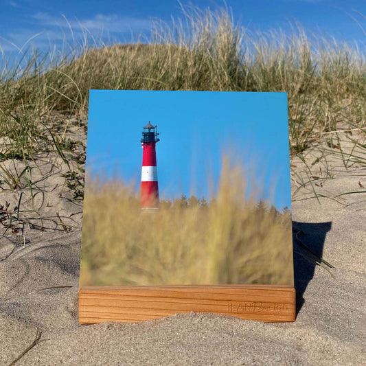 Acrylbild in einem Holzstandbildhalter steht in den Sylter Dünnen. Auf dem Acrylbild ist der Hörnumer Leuchtturm mit blauem Himmel zu sehen. Im Vordergrund sieht man etwas verschwommen Dünengras.