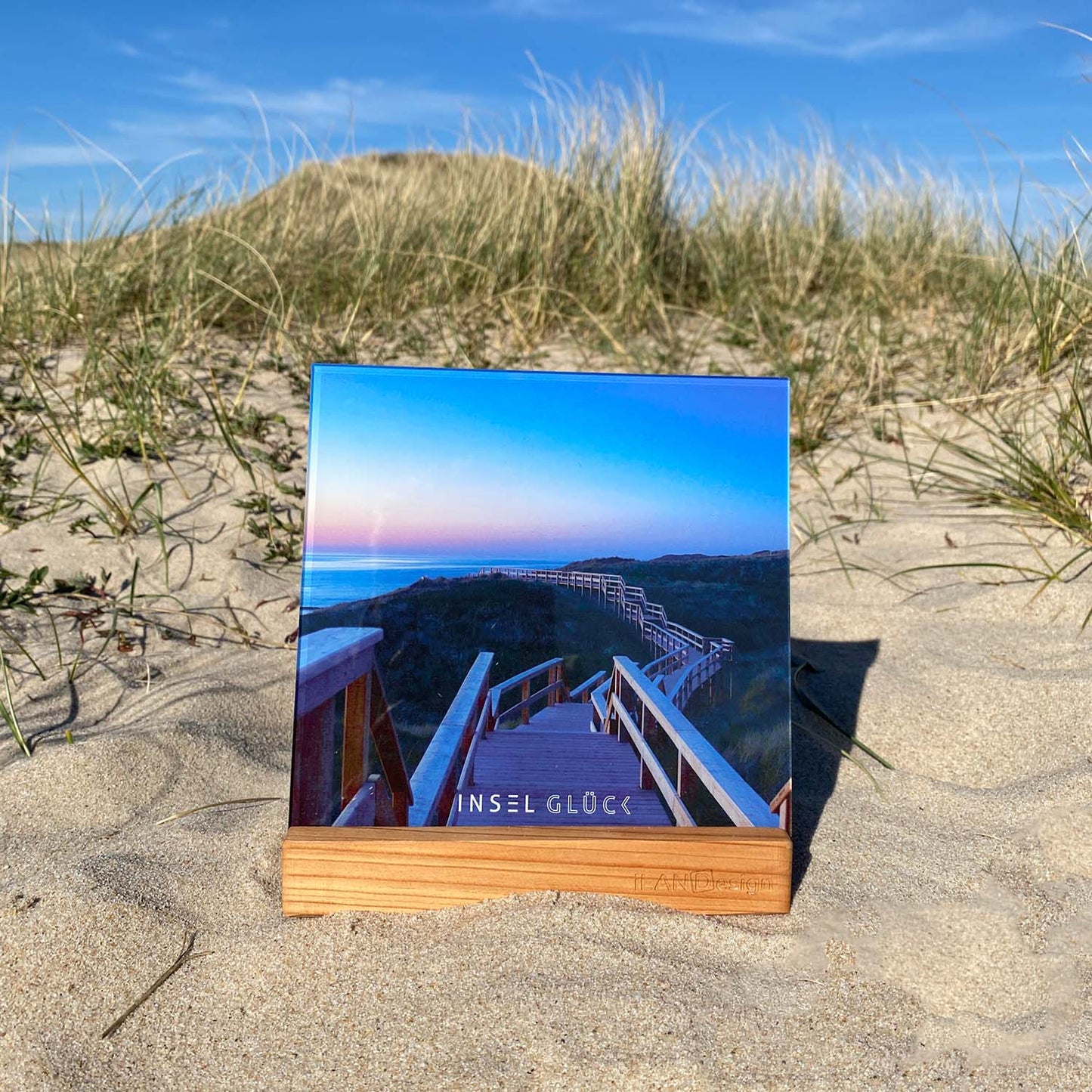 Acrylbild mit Steg durch die Dünen steht in einem Holzbilderhalter in den Sylter Dünen. Unten mittig ist der Begriff Inselglück aufgedruckt.