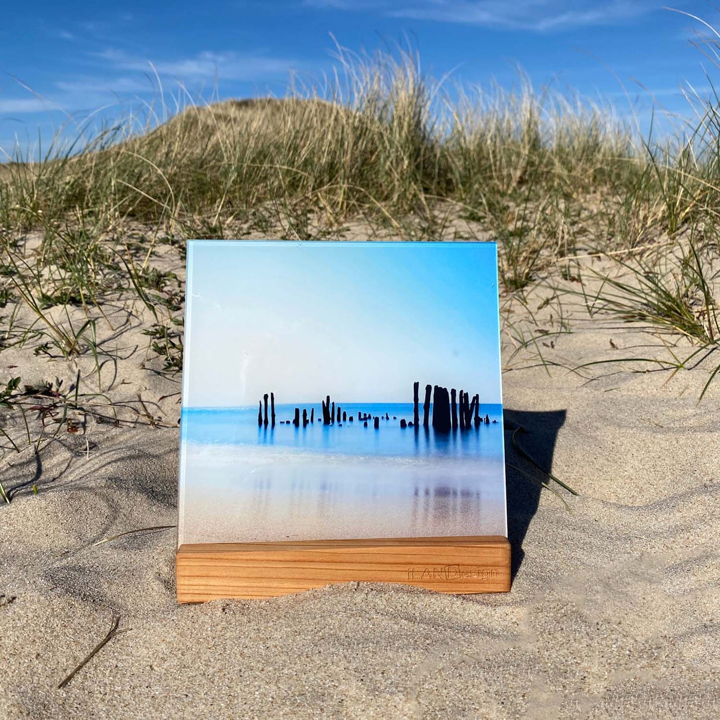 Acrylbild mit Buhnenresten im Meer steht in einem Holzbilderhalter in den Sylter Dünen.