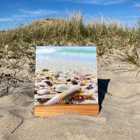 Acrylbild mit Muscheln und im Hintergrund die Nordsee steht in einem Holzbilderhalter. Mittig unten ist der begriff Inselglück aufgedruckt. Das Set steht in den Sylter Dünen.