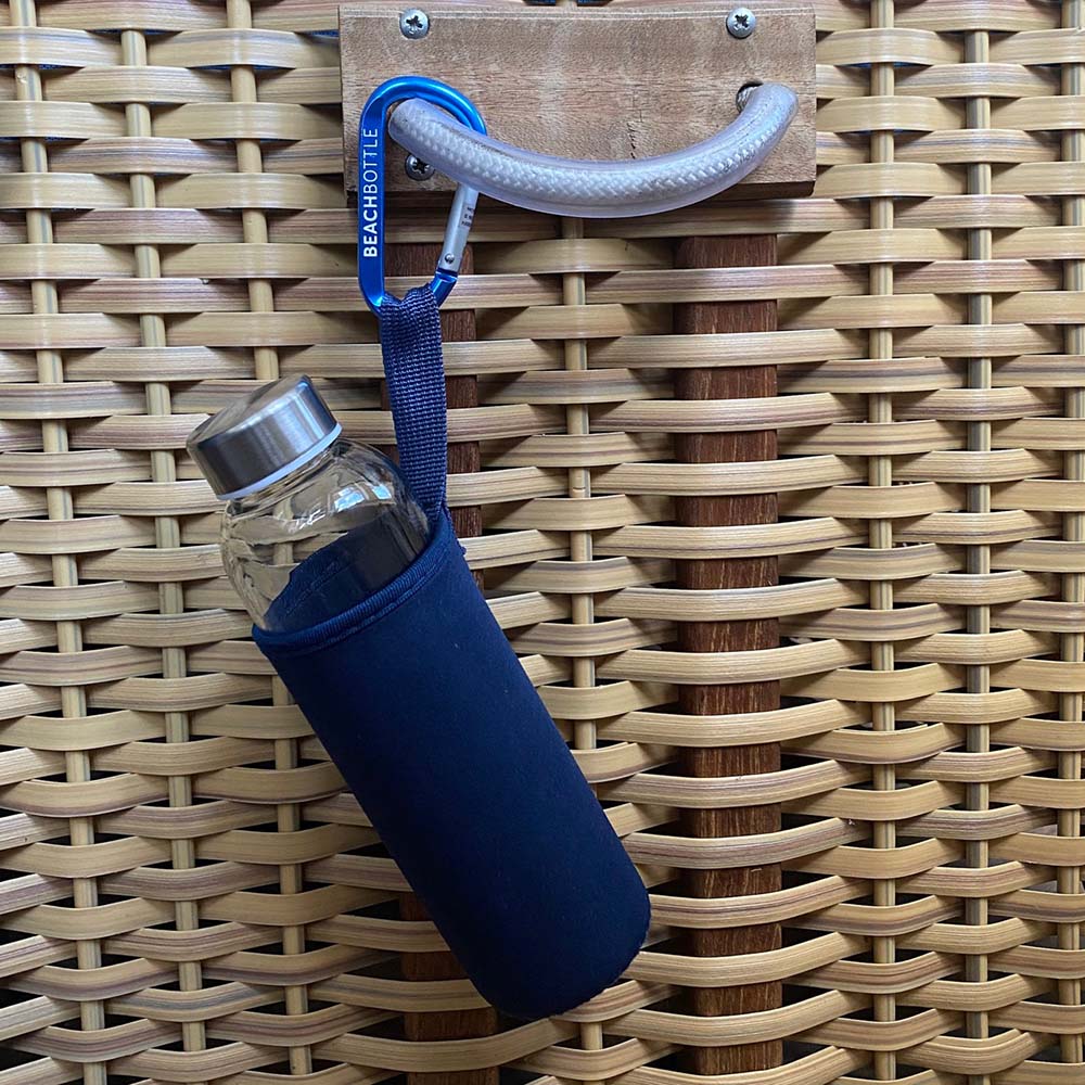 Nachhaltige Glasflasche in dunkelblauer Neoprenhülle hängt an einem großen, blauen Karabiner mit dem Schriftzug Beachbottle eingraviert, an einem Strandkorbgriff.