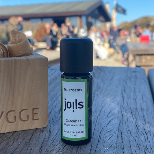 Kleines Duftöl-Fläschen steht auf einem Holztisch mit Blick auf eine Sylter Strandbude. Auf dem Etikett der 100% naturreinen Ölmischung steht die Duftrichtung Sansibar.