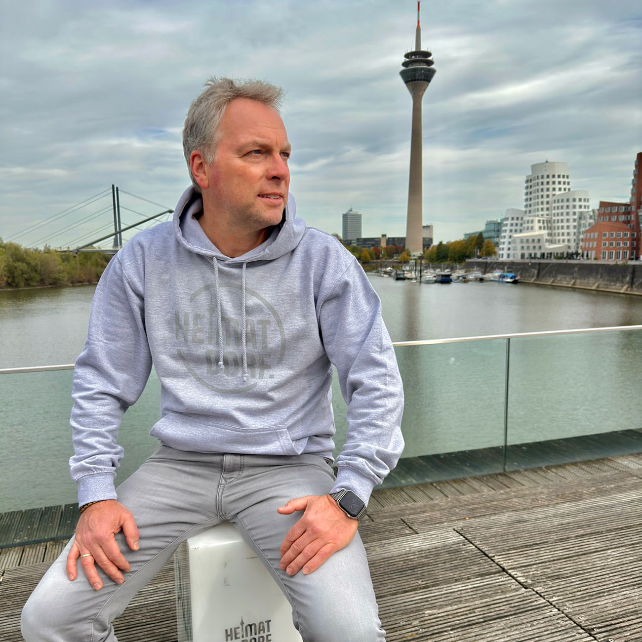 Mann sitzt am Düsseldorfer MedienHafen mit Blick auf den Rheinturm und die Gehry Häuser. Der graue Hoodie hat einen mittelgrauen, großen, runden Aufdruck mit dem Logo-Schriftzug HeimatDorf.