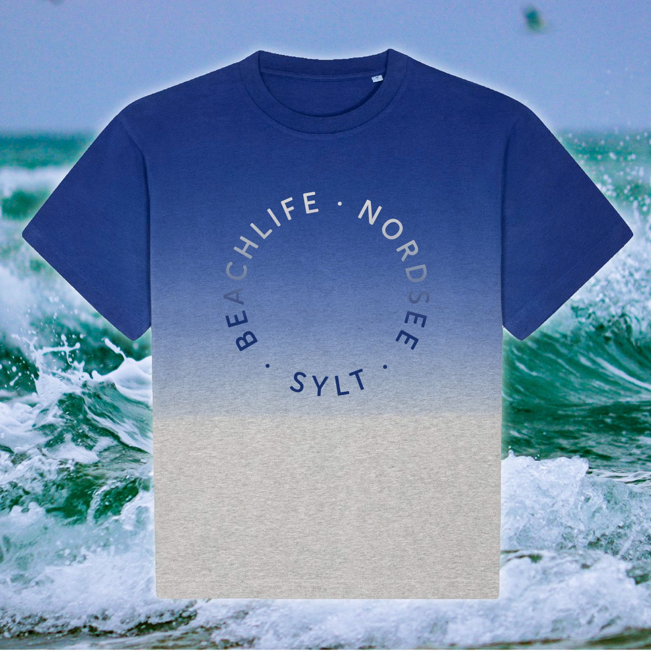 T-Shirt mit Farbverlauf und Sylt-Aufdruck. Das Shirt ist am Rundhalsausschnitt in einem blau-hellgrau gefärbt und läuft in ein beige aus. In der gleichen, aber umgekehrten Farbe steht in rund der Schriftzug Beachlife, Nordsee, Sylt. Im Hintergrund sieht man einen Detailausschnitt der Nordsee-Wellen.