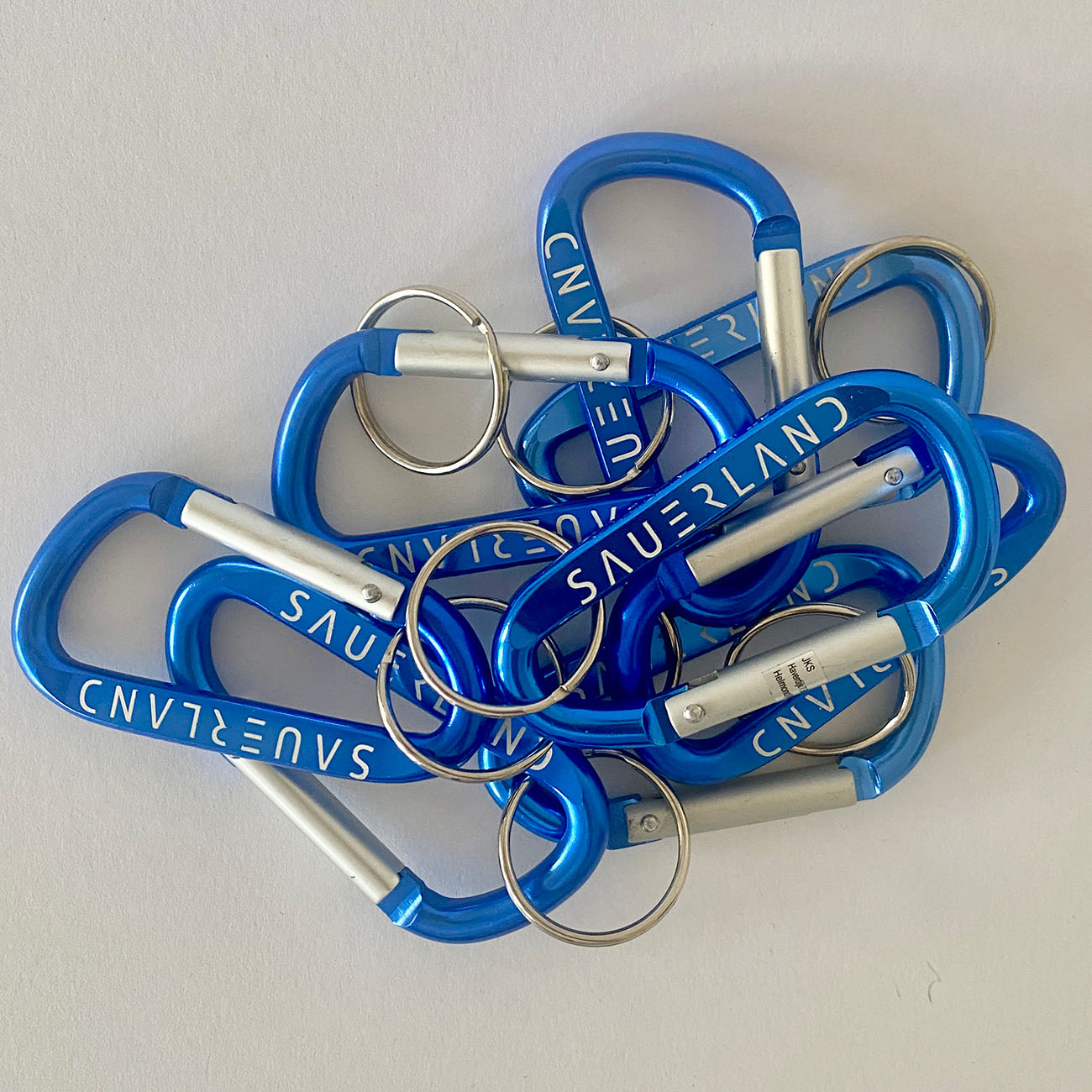 Auf diesem Foto liegen viele mittelblaue Karabiner-Schlüsselanhänger auf einem weißen Untergrund. Jeder Karabiner hat eine einseitige Lasergravur mit dem new Sauerland Schriftzug.