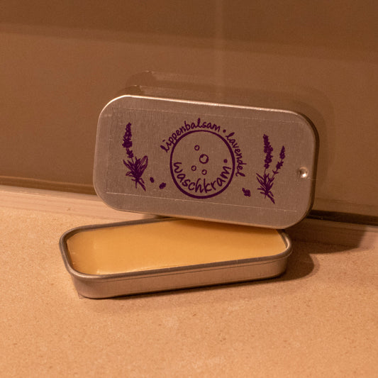 Eine kleine Weißblech-Schiebedose liegt geöffnet auf einer Badezimmerablage. Die Dose enthält einen biologischen Lippenbalsam mit Lavendelduft.